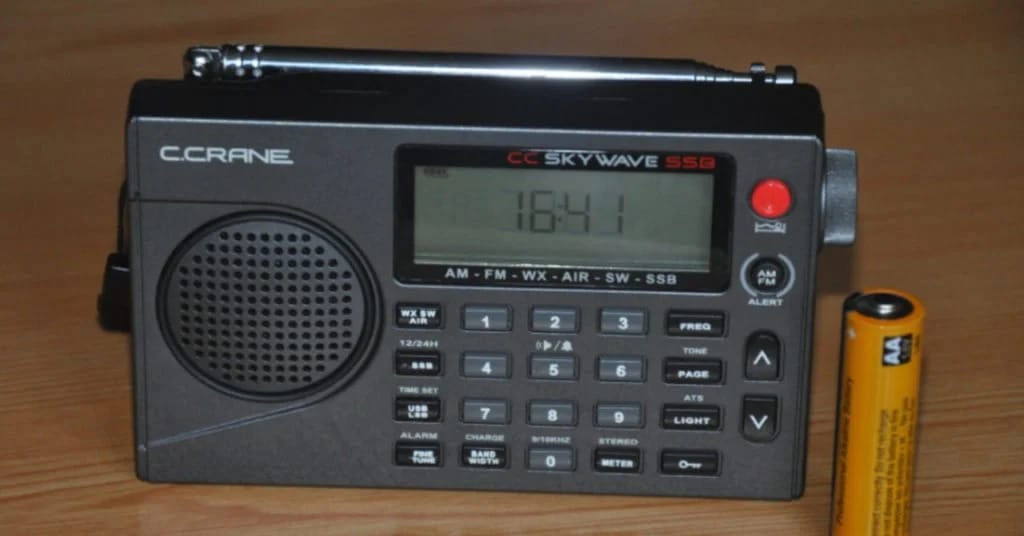 A C.Crane Skywave SSB radio displaying 16:41 on its digital screen, placed on a wooden surface with a battery beside it.