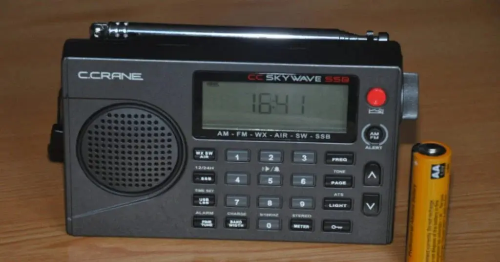 A C.Crane Skywave SSB radio displaying 16:41 on its digital screen, placed on a wooden surface with a battery beside it.