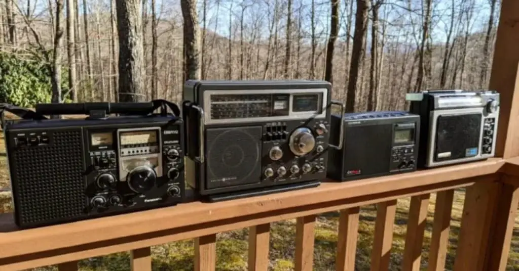 A lineup of vintage radios, including a C.Crane radio, placed on a wooden deck with a forest background.
