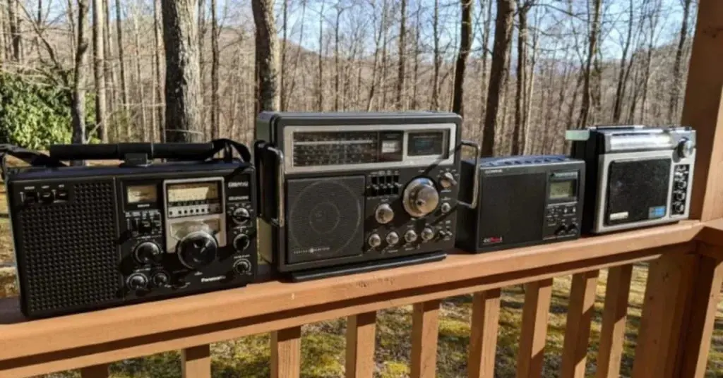 A lineup of vintage radios, including a C.Crane radio, placed on a wooden deck with a forest background.